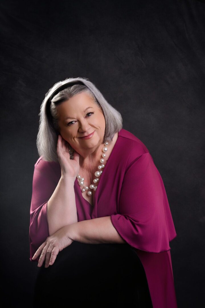 Portrait of a confident older woman in a magenta top against a dark background.
